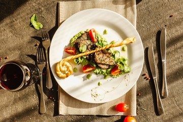 Traditional German fried pork cheeks with greens and tomato closeup on a white plate with bread cakes on a served table with a glass of red wine