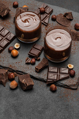 Wall Mural - glass bowl of chocolate cream or melted chocolate, pieces of chocolate and hazelnuts on dark concrete background