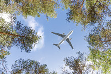 Travel, Transportation, Aviation Concept. Wide-body Passenger Airplane Flying Overhead Against A Summer Blue Sky Between The Trees, Bottom View. 
