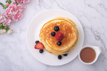 Pancakes on white plate topped with strawberry, rasberry and black currants and honey syrup and pink blooming flower in blackground.