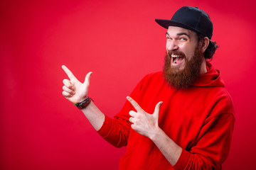 Wall Mural - Excited bearded guy in red hoody and black hat, poitning with finger at copy space over red background