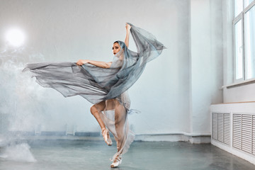 Full-size expressive photo of female ballet dancer dressed in grey-blue flowing fabric spinning on one leg on white y background.