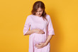 © sementsova321 - Image of beautiful young pregnant woman isolated against yellow studio background, adorable lady stands with hands on her abdomen, looks down on her belly, imagines her future baby. Motherhood concept