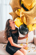 © golubovy - Special day. Amused brunette girl sitting on sofa in black sequin mini dress, laughing, holding bunch of golden balloons.