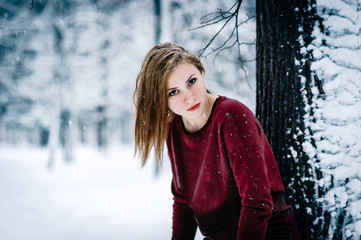 Wall Mural - the girl dressed in a maroon sweater and white pants leaned against the tree trunk against a backdrop of snow-covered winter forest