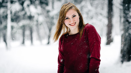 Wall Mural - the girl dressed in a maroon sweater stands against the tree trunk against a backdrop of snow-covered winter forest