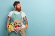 Red haired young man overstained with housekeeping, holds basin with pile of laundry, wears casual t shirt and apron, looks away, isolated over blue background, has thoughtful look, focused away
