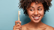 © WHstudio Leushin N - Teeth care and oral hygiene concept. Positive young Afro American woman with curly haircut, holds wooden toothbrush, cleans teeth, has dark skin, being in high spirit. Healthcare and dentistry