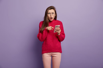 Sticker - Indignant shocked young woman points at cell phone, demonstrates something amazing, surprised with received message, wears red long sleeved sweater, has astonished facial look, connected to wifi