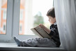© alexkich - The little boy is reading a book. The child sits at the window and prepares for lessons. A boy with a book in his hands is sitting on the windowsill.