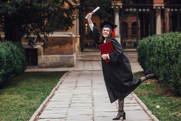 Portrait of female college student in graduation cap and gown holding diploma on campus and smiling! Lifestyle!