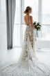 Gorgeous bride in a luxurious long dress with a bouquet in hand, stands and looks out the window