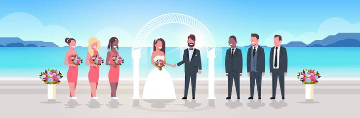 Wall Mural - just married bride and groom with bridesmaids groomsmen standing together on sea beach near arch wedding ceremony concept sunrise mountains background full length horizontal