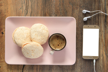 Smartphone with connected earphones and isolated blank white screens for design and cake macaroon cup of coffee on a rectangular plate on the background of a desk