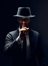 Serious Man In Black Suit And Hat Keeps Finger On Lips, Making Hush Gesture And Keep Conspiracy Isolated On Dark Background