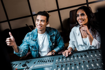 Canvas Print - handsome multicultural sound producer showing thumb up while sitting near smiling attractive colleague