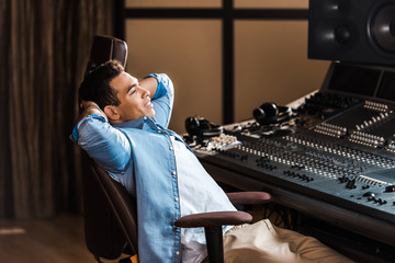 Wall Mural - handsome smiling mixed race sound producer resting in office chair in recording studio