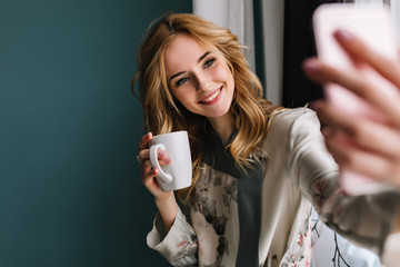 pretty young woman with wavy blonde hair taking selfie sitting next to the window with cup of mornin