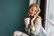 © Look! - Cheerful blonde woman relaxing and sitting on window sill, holding cup of coffee, tea. She has long blonde wavy hair, beautiful smile. Wearing nice pajama in flowers.