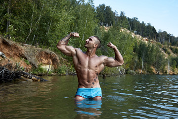 Tattooed trendy guy shows off his perfect muscles in water drops standing in the lake after bathing