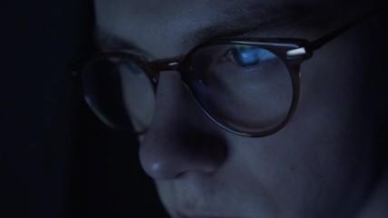 Very tired nerdy student with glasses is sitting in the darkness browsing the internet.