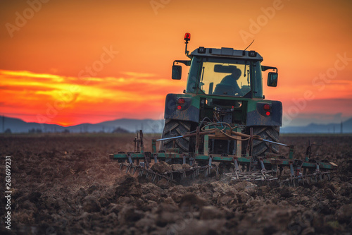 Cuadros en Lienzo Beautiful sunset, farmer in tractor preparing land with seedbed cultivator