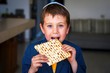 Cute Caucasian child in a yarmulke taking a bite from a traditional Jewish matzo unleavened bread in a room.