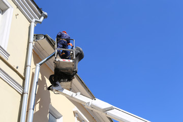 Workers in hydraulic lifting ramp repair the roof gutter. Builders on lifting boom platform standing near the building wall, construction and repair works