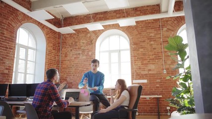 Wall Mural - Young startup business team of three discussing application in office with brick walls. Project manager looking at notes and explaining ideas to colleagues, programmer coding on laptop computer