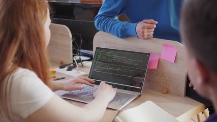 Wall Mural - Young female software developer with red hair typing code on laptop computer while her male colleagues giving pieces of advice, tracking left medium shot