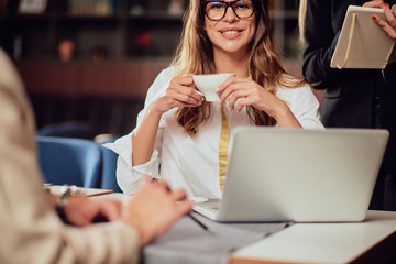 Gorgeous Caucasain businesswoman with long brown hair and dressed elegant looking at camera and holding cup of coffee. Her colleagues sitting and standing around her.