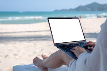 Mockup image of a woman holding and using laptop computer with blank desktop screen while laying down on beach chair on the beach