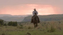 A Lone Rancher On A Trotting Though The Valley On His Horse During A Pastel Pink Sunset With A Mountain Range On The Horizon, Slow Motion 24 Fps.