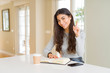 © Krakenimages.com - Young woman reading a book and drinking a coffe surprised with an idea or question pointing finger with happy face, number one