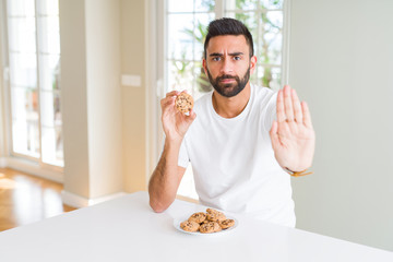 Poster - Handsome hispanic man eating chocolate chips cookies with open hand doing stop sign with serious and confident expression, defense gesture