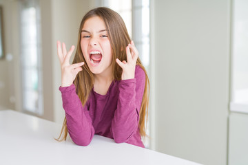 Wall Mural - Beautiful young girl kid on white table celebrating mad and crazy for success with arms raised and closed eyes screaming excited. Winner concept