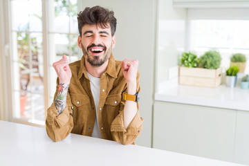 Young man wearing casual jacket sitting on white table excited for success with arms raised celebrating victory smiling. Winner concept.