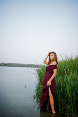 Wall Mural - Blonde sensual woman in red marsala dress standing in water of lake with reeds.