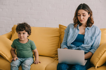 Wall Mural - Cute kid sitting on sofa while mother using laptop