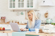© oes - Freelance concept. Beautiful business woman in casual clothes and glasses is examining documents and smiling while working with a laptop in kitchen. Working at home