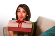 © TheVisualsYouNeed - young beautiful and happy Asian Indonesian woman holding birthday or Christmas present showing the gift box cheerful and excited sitting on living room couch