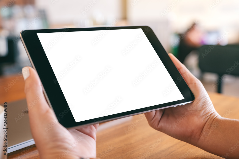 Mockup image of hands holding black tablet pc with blank white screen with laptop and coffee cup on wooden table - obrazy, fototapety, plakaty 