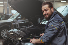 Cheerful Bearded Mechanic Smiling Over His Shoulder To The Camera, While Repairing An SUV Car At The Garage, Copy Space. Friendly Auto Technician Enjoying Working With Automobiles