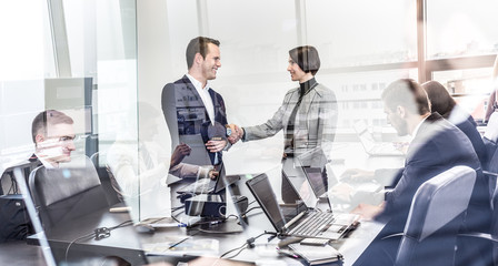 Wall Mural - Sealing a deal. Business people shaking hands, finishing up meeting in corporate office. Businessmen working on laptop seen in glass reflection. Business and entrepreneurship concept.