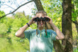 © Andrii Oleksiienko - Horizontal color photography of cute white funny little kid looks through old vintage black binoculars at camera smiling  while standing at scenic blurry green fresh foliage of spring trees background