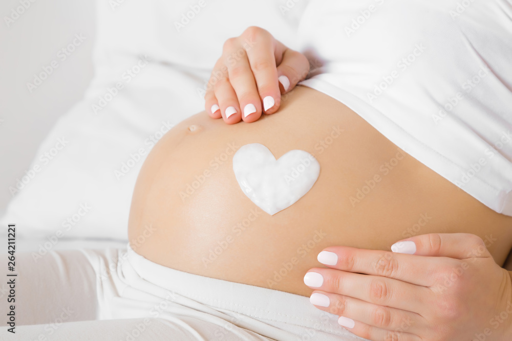 Girl sitting in bed. Young woman's hands touching her naked big belly. Heart shape created from cream. Care about perfect, soft and smooth skin in pregnancy time. Close up. Side view.  - obrazy, fototapety, plakaty 