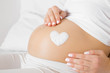 Girl sitting in bed. Young woman's hands touching her naked big belly. Heart shape created from cream. Care about perfect, soft and smooth skin in pregnancy time. Close up. Side view. 