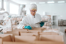 Young Caucasian employee in sterile uniform holding tablet and counting products in boxes. Food factory interior.