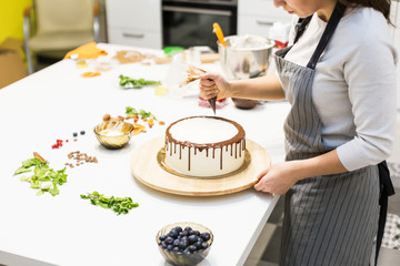 Wall Mural - A confectioner squeezes liquid chocolate from a pastry bag onto a white cream biscuit cake on a wooden stand. The concept of homemade pastry, cooking cakes.