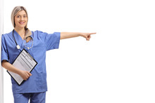 Young Female Nurse With A Clipboard Leaning Against A Wall And Pointing